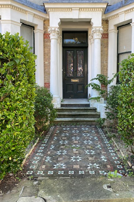 A narrow garden pathway leading up to the black front door of a Victorian terrace house with the number 8 displayed above. The pathway is decorated with intricate patterned tiles featuring star and diamond motifs in shades of gray, white, and black. On either side of the path, there are well-maintained, lush green hedges and small bushes, creating a tidy border. The steps leading to the entrance are made of stone, and the doorway is framed by white columns with decorative capitals, supporting a small pediment. The house's exterior walls are a combination of brick and cream-colored stucco, with large sash windows displaying dark frames. The scene is well-lit with natural daylight, emphasizing the contrast between the greenery and the detailed paving, encapsulating charming Victorian terrace garden styling typical of E3 properties, and exemplifying garden maintenance and landscaping work by Gardeners Bow.