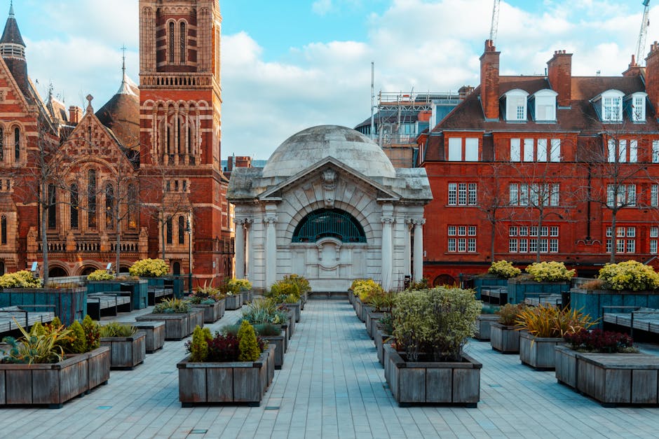 A rooftop garden space situated on a terrace with a view of historic red brick Victorian buildings, including a church with a tall steeple and an ornate tower. The garden features a paved central pathway bordered by rectangular wooden planters filled with a variety of plants, including small shrubs, flowering perennials, and ornamental grasses, providing textures of lush green, deep red, and yellow foliage. At the far end of the terrace, there is a small domed structure with white stonework and an arched window, surrounded by neatly trimmed hedges and additional planter arrangements. The garden benches and modern seating areas are integrated into the layout, designed for outdoor relaxation and socialising. The environment is bathed in natural daylight with partly cloudy skies overhead, illustrating a scene of gardening and outdoor maintenance typical of an urban Victorian terrace setting, with a focus on landscape design and cityscape views. As seen, Gardeners Bow provides professional gardening and landscaping work to maintain such well-kept outdoor spaces.