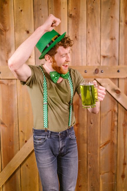 A man with curly red hair and a beard, dressed in casual gardening attire, is standing indoors against a wooden wall. He is wearing a green hat, a green T-shirt, and green suspenders with white polka dots, complemented by a matching green bow tie. He is smiling broadly, raising a fist in a celebratory gesture, and holding a large glass mug filled with a bright yellow-green beverage, possibly a garden-inspired drink. The wooden wall behind him features visible grain and knots, with some planks arranged vertically. The lighting is warm, creating a cheerful atmosphere, and the scene suggests a relaxed, festive moment related to outdoor or garden activities, aligning with themes of outdoor enjoyment and leisure. This image exemplifies lighthearted outdoor celebration, often associated with gardening or outdoor events, as promoted by Gardeners Bow.