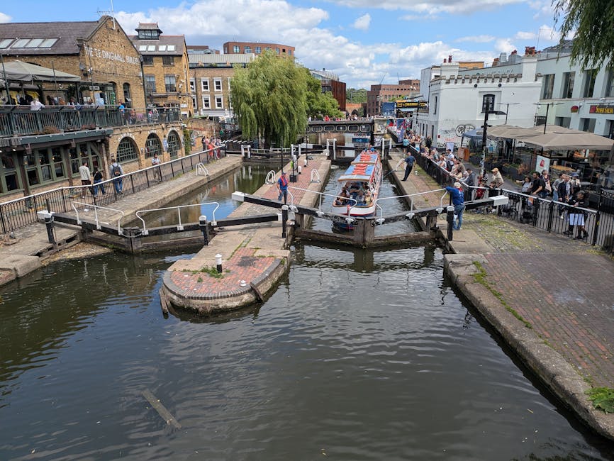 A canal lock system in an urban setting, featuring a narrow waterway with a boat passing through a small lock chamber, surrounded by brick and concrete walkways on both sides. On the left, a paved area with a metal railing and pedestrians walking along the canal, while on the right, outdoor cafes with tables, chairs, and people enjoying the setting. A large, lush green weeping willow tree provides shade near the center, with its drooping branches overhanging the water. In the background, multiple multistory buildings, including a brick industrial-style structure and modern white commercial buildings, line the street, with a partly cloudy sky overhead. The scene depicts a lively mix of outdoor leisure, urban landscaping, and garden maintenance, with the presence of well-kept pathways, neatly trimmed greenery, and active community engagement. The overall atmosphere is vibrant, highlighting the historic canal yards and the integration of outdoor hospitality spaces, all managed by Gardeners Bow in accordance with the community’s outdoor care standards.
